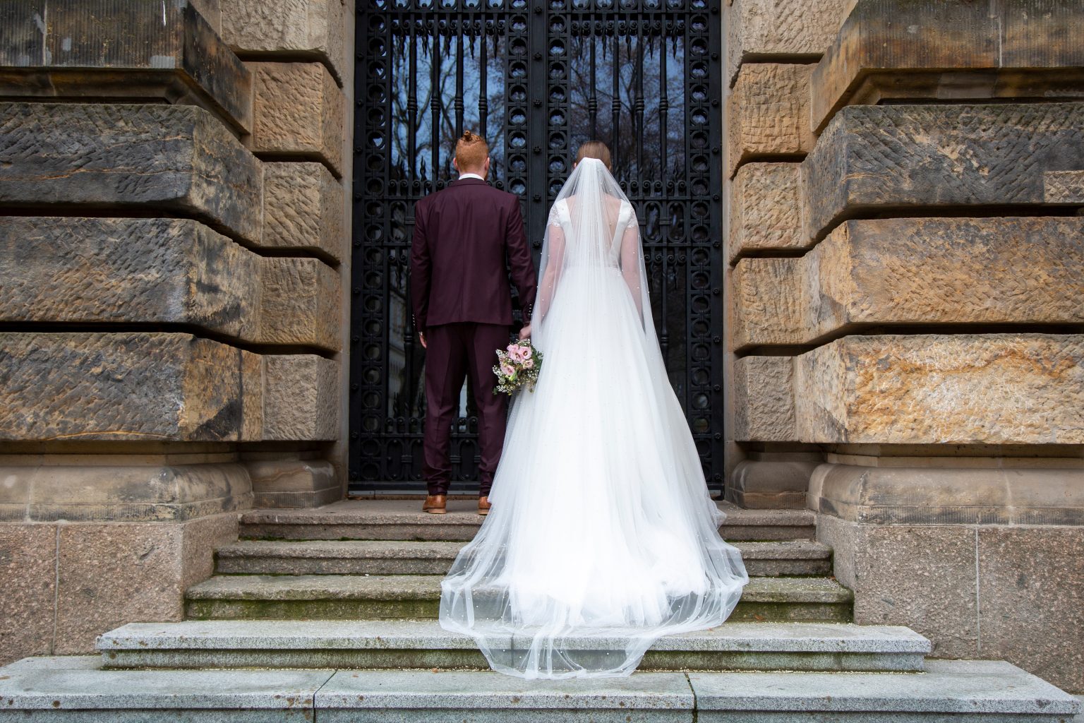 Hochzeitsfoto von Sarah Seiler Fotografie Eine Braut und ein Bräutigam stehen vor großen Türen und Sarah Seiler Fotografie fotografiert sie.