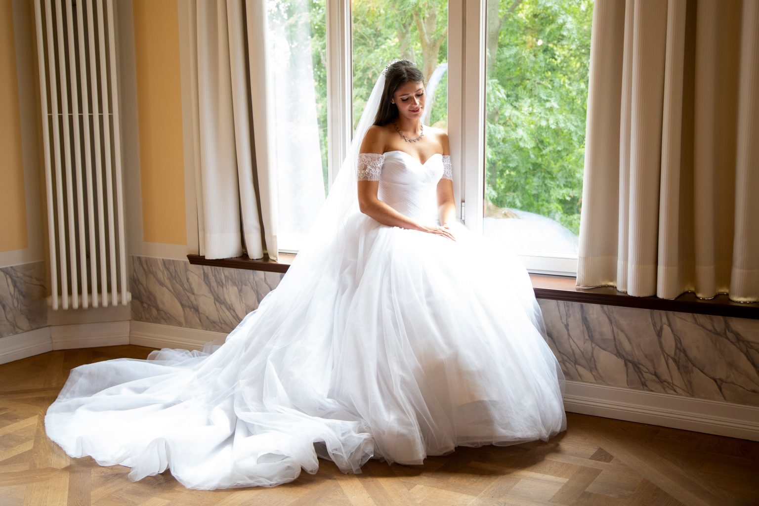 Sarah Seiler Fotografie Eine Braut sitzt vor einem großen Fenster und Sarah Seiler Fotografie hält diesen ruhigen Moment fest.