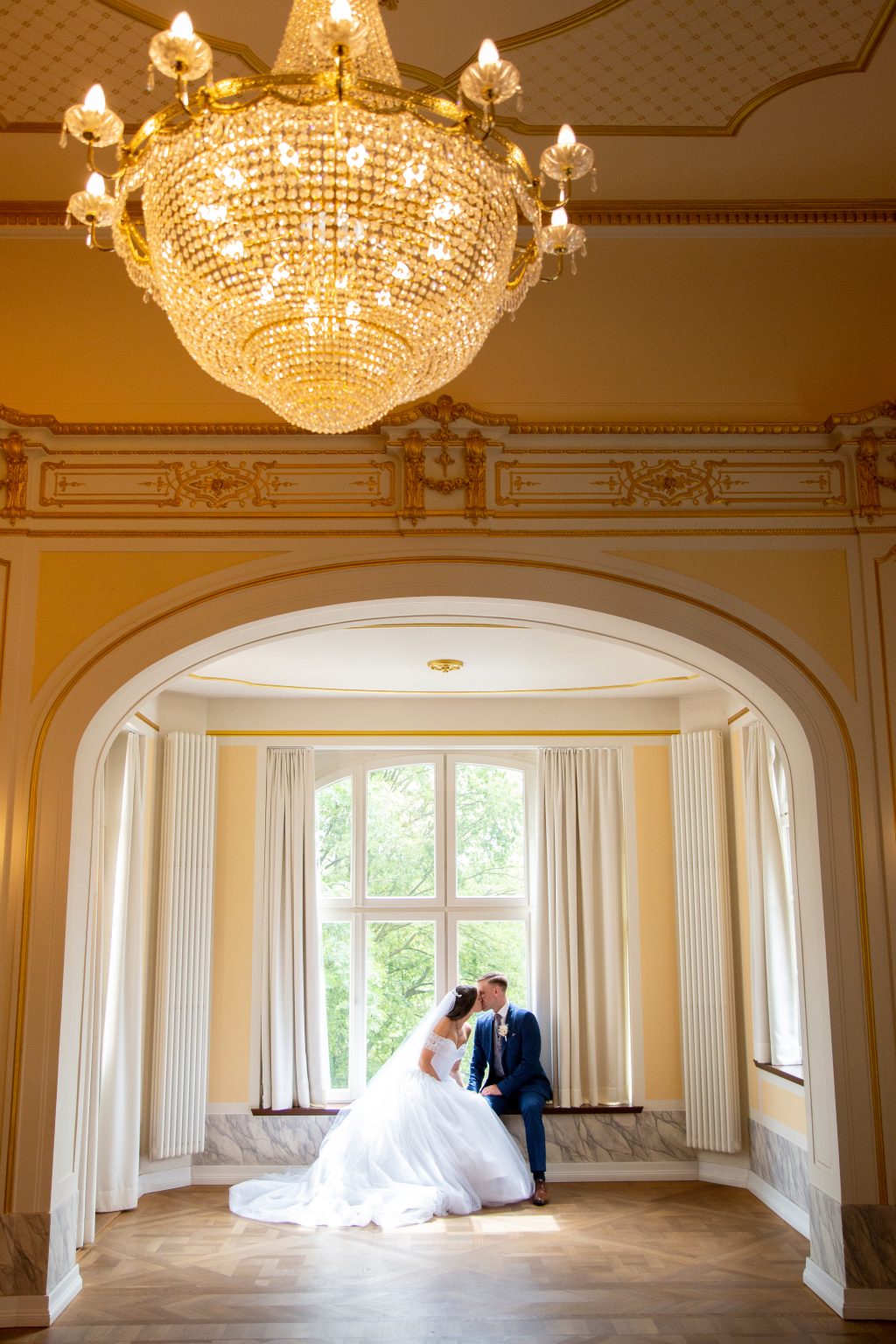 Sarah Seiler Fotografie Ein Hochzeitspaar sitzt unter einem großen Kronleuchter und Sarah Seiler Fotografie fotografiert sie.