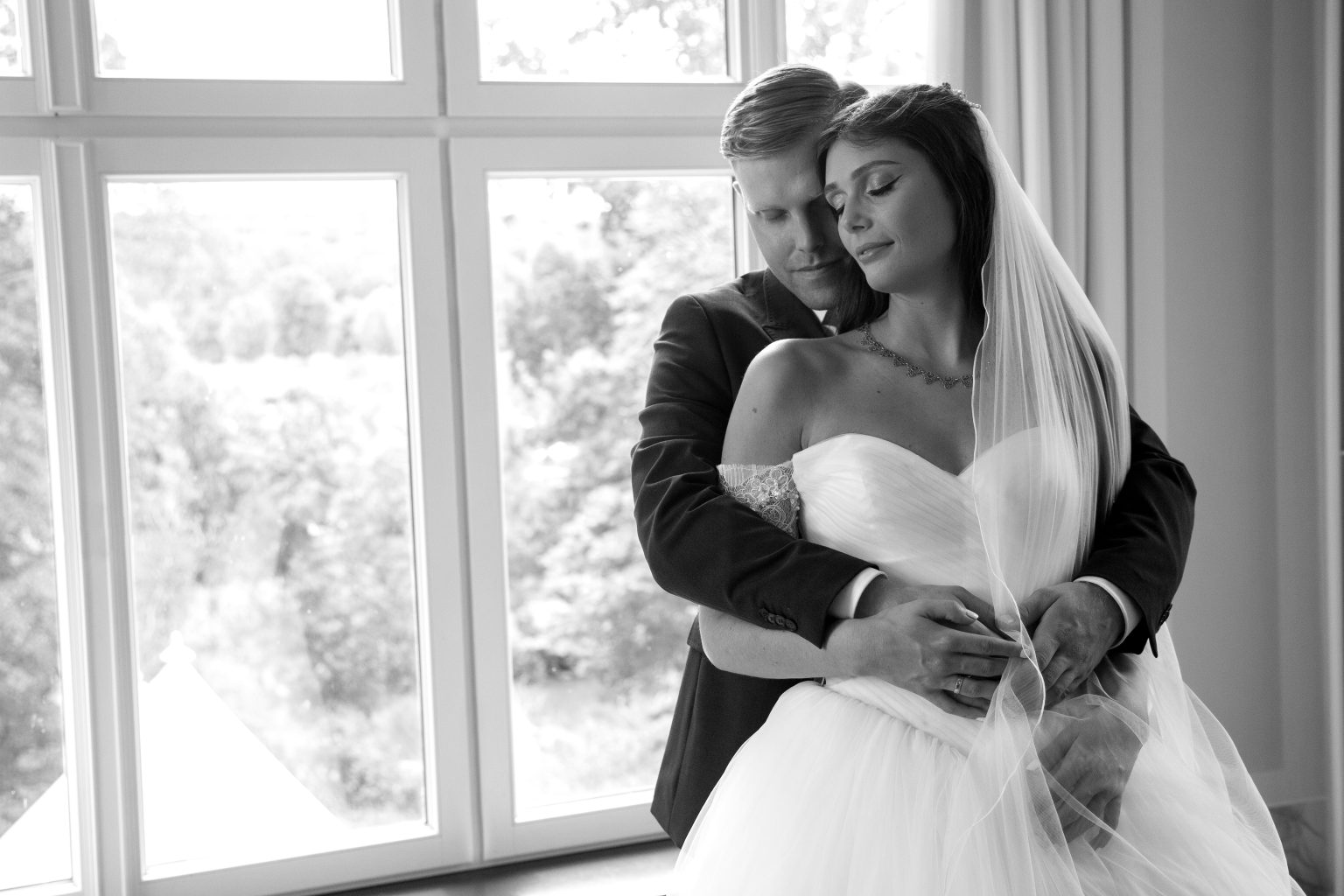 Hochzeitsfoto von Sarah Seiler Fotografie Ein Hochzeitspaar steht umarmt vor einem großen Fenster und Sarah Seiler Fotografie hält diesen emotionalen Moment fest.