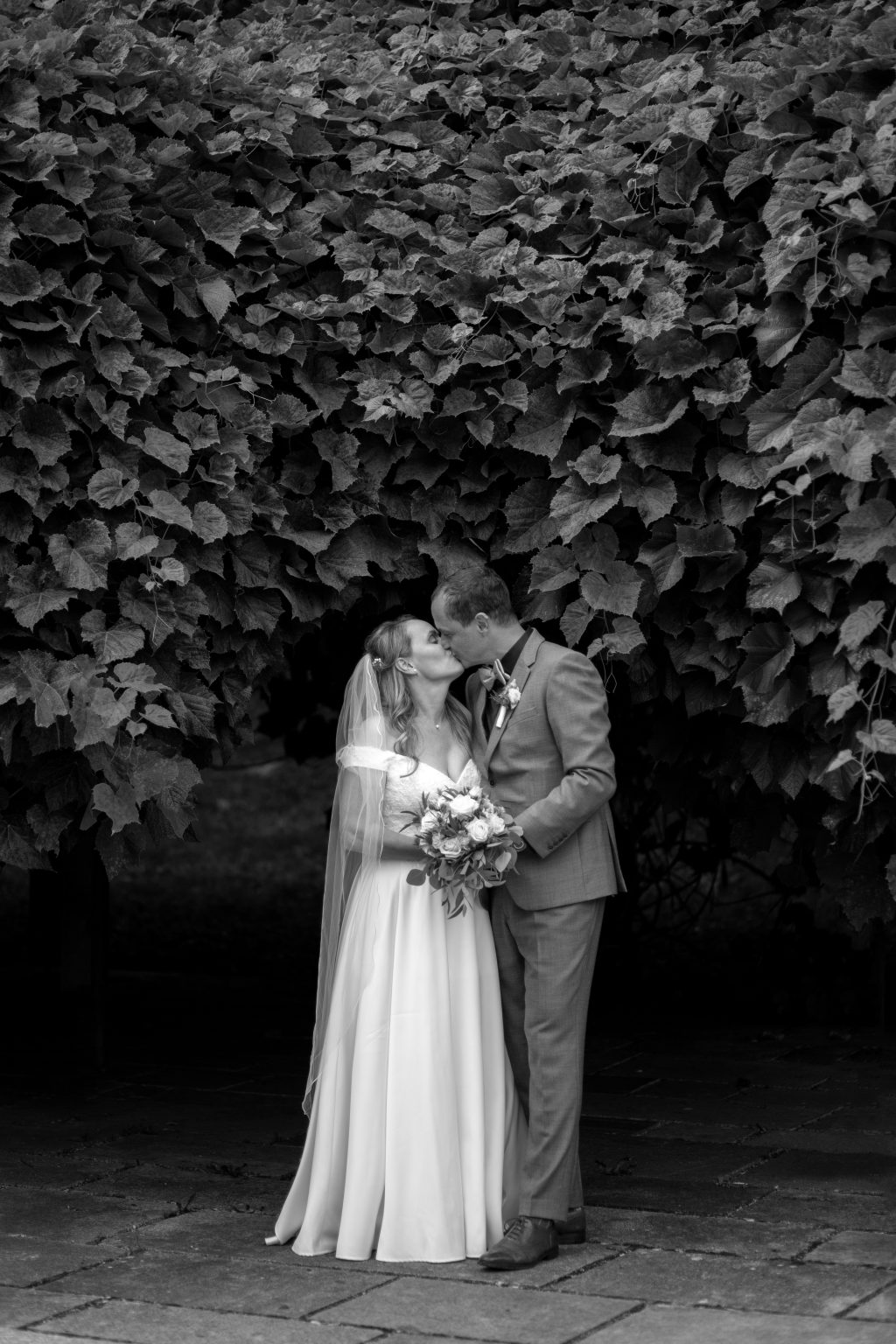 Hochzeit_Jana_und_Konrad-287SW Ein Hochzeitspaar steht eng umarmt vor einer Blätterwand und Sarah Seiler Fotografie hält diesen Moment fest.