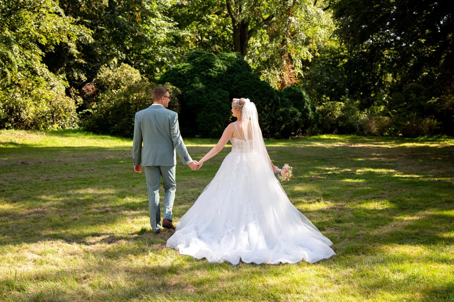 Hochzeitsfoto von Sarah Seiler Fotografie Ein Hochzeitspaar geht Hand in Hand über eine Wiese und Sarah Seiler Fotografie fotografiert sie.