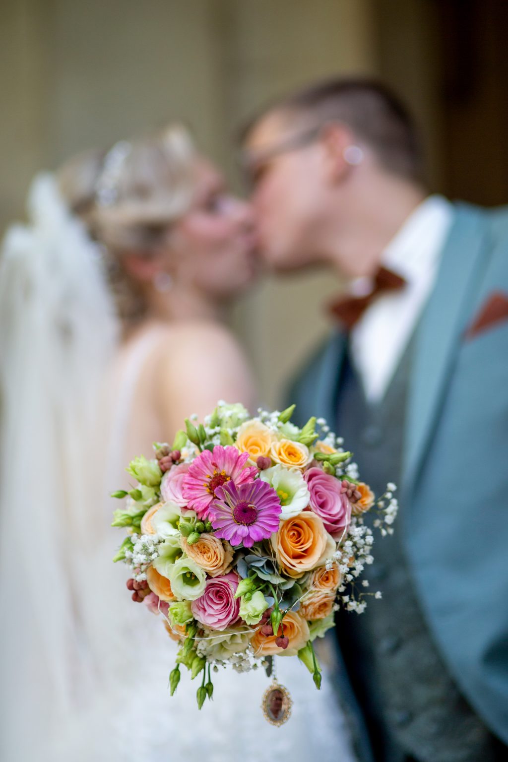 Hochzeitsfoto von Sarah Seiler Fotografie Ein Brautstrauß steht im Vordergrund, während ein Hochzeitspaar im Hintergrund küsst, und Sarah Seiler Fotografie fotografiert diesen Moment.